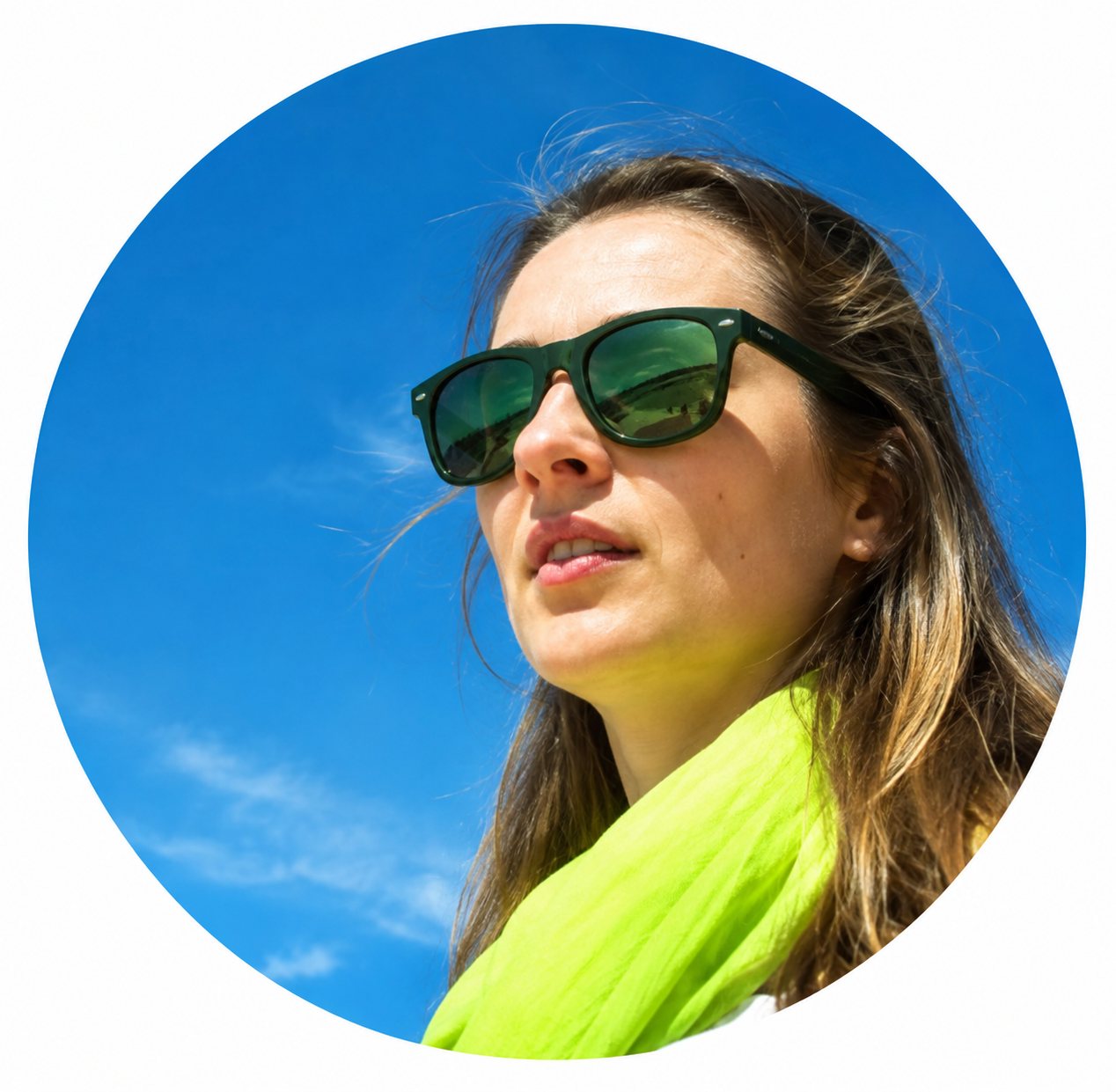 Une femme aux longs cheveux portant des lunettes de soleil sombres et une écharpe jaune pose en plein air devant un ciel bleu.
