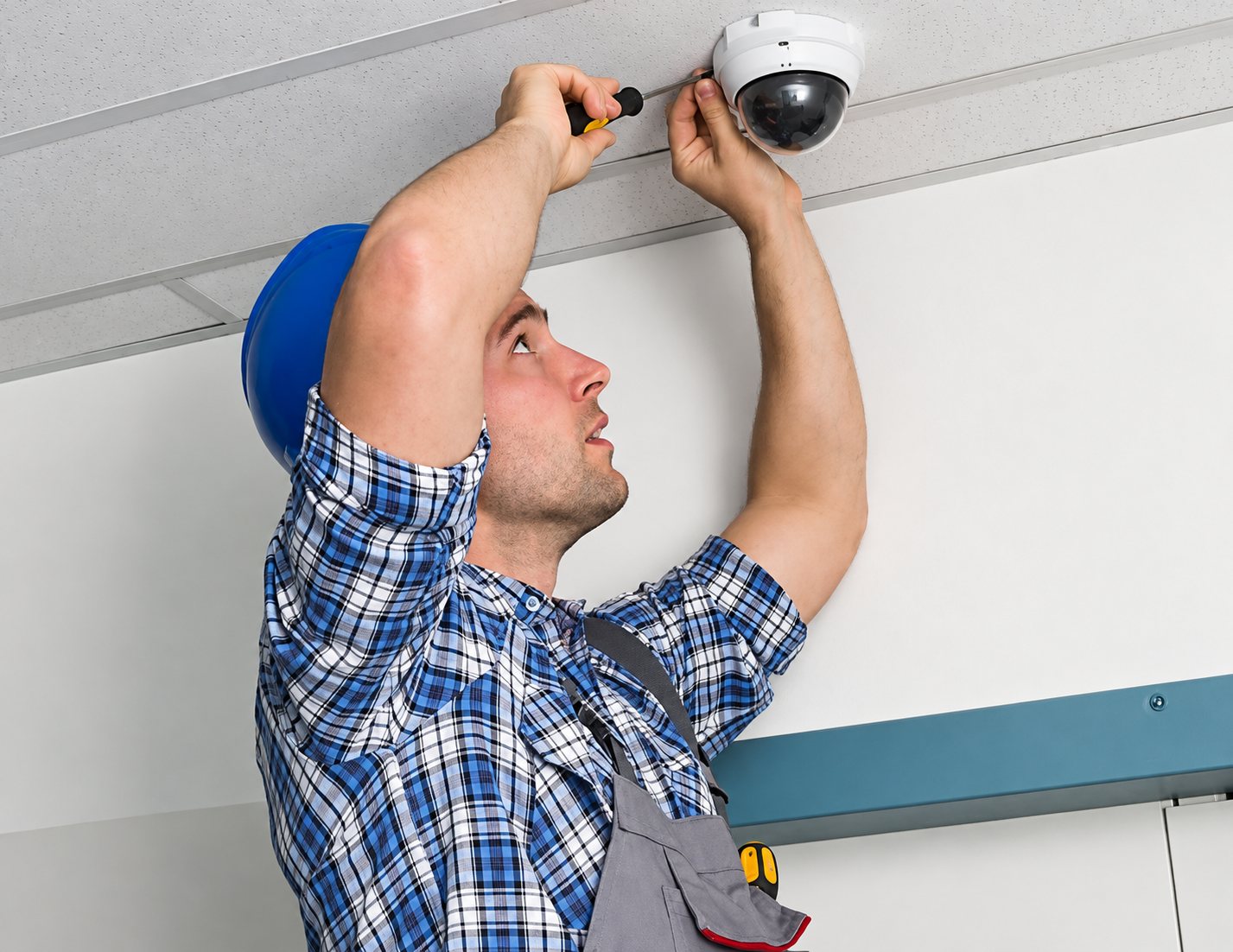 Un technicien en chemise à carreaux et casque bleu visse une caméra de surveillance dôme fixée au plafond d’une pièce.