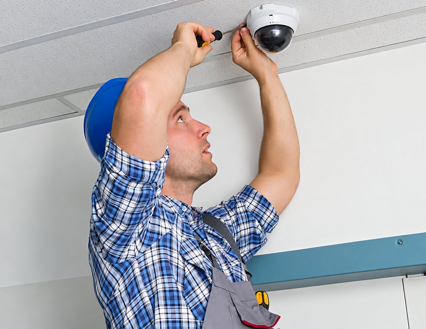 Un technicien en chemise à carreaux et casque bleu fixe avec un tournevis une caméra de vidéosurveillance dôme au plafond.